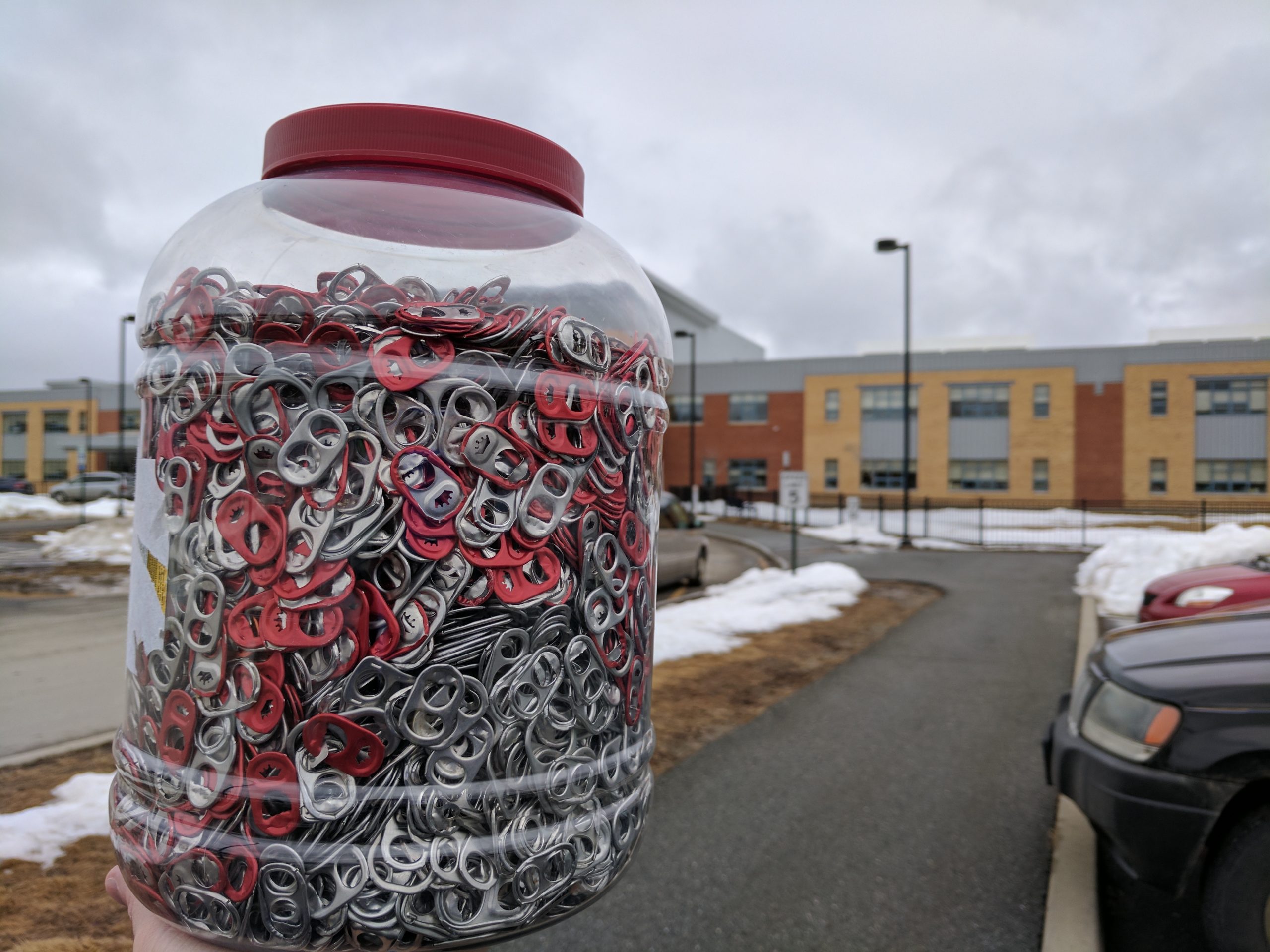 A hand holds a large jar filled with colorful soda can tabs outside a school on a cloudy day.