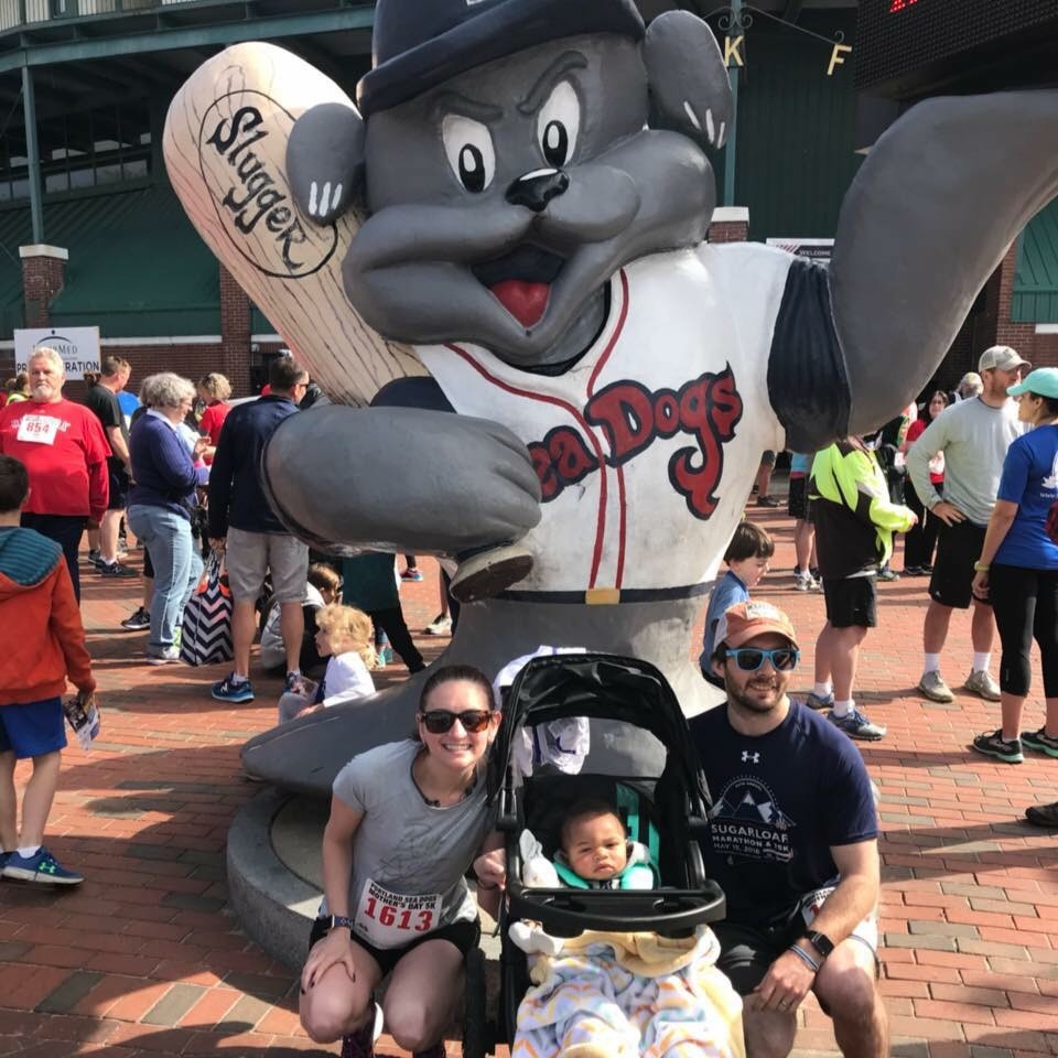 A family poses in front of a large mascot statue at a busy outdoor event, with runners and spectators in the background.