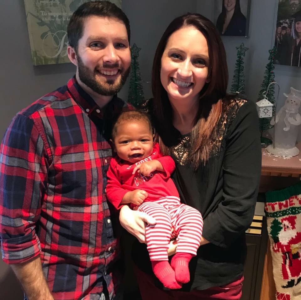 A smiling couple holds a happy baby dressed in red and striped pajamas, with festive decorations in the background.