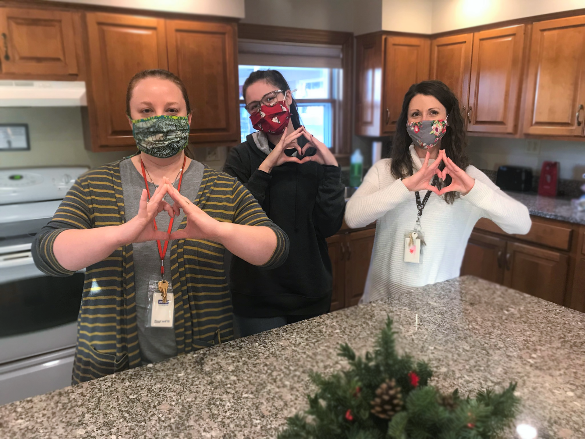 Three masked women stand in a kitchen, forming heart shapes with their hands, smiling at the camera.