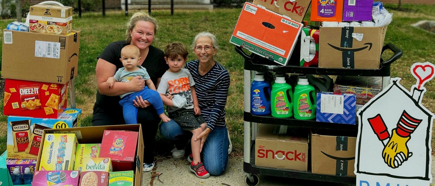 A woman holds a baby, while another woman and a boy pose beside a cart filled with donations for a charity.