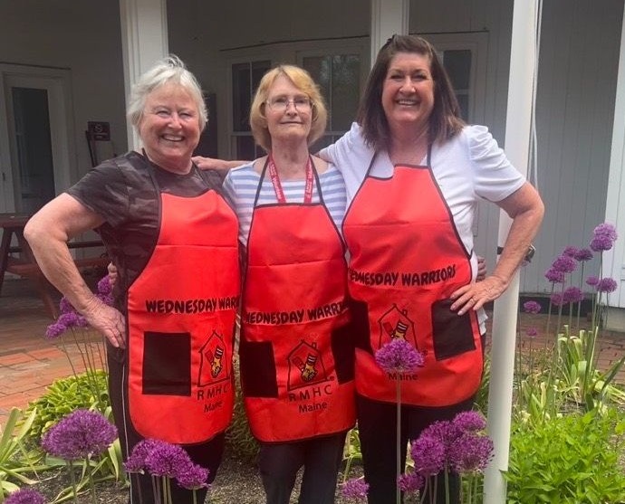 Three women in "Wednesday Warriors" aprons smile together outdoors, surrounded by blooming flowers.