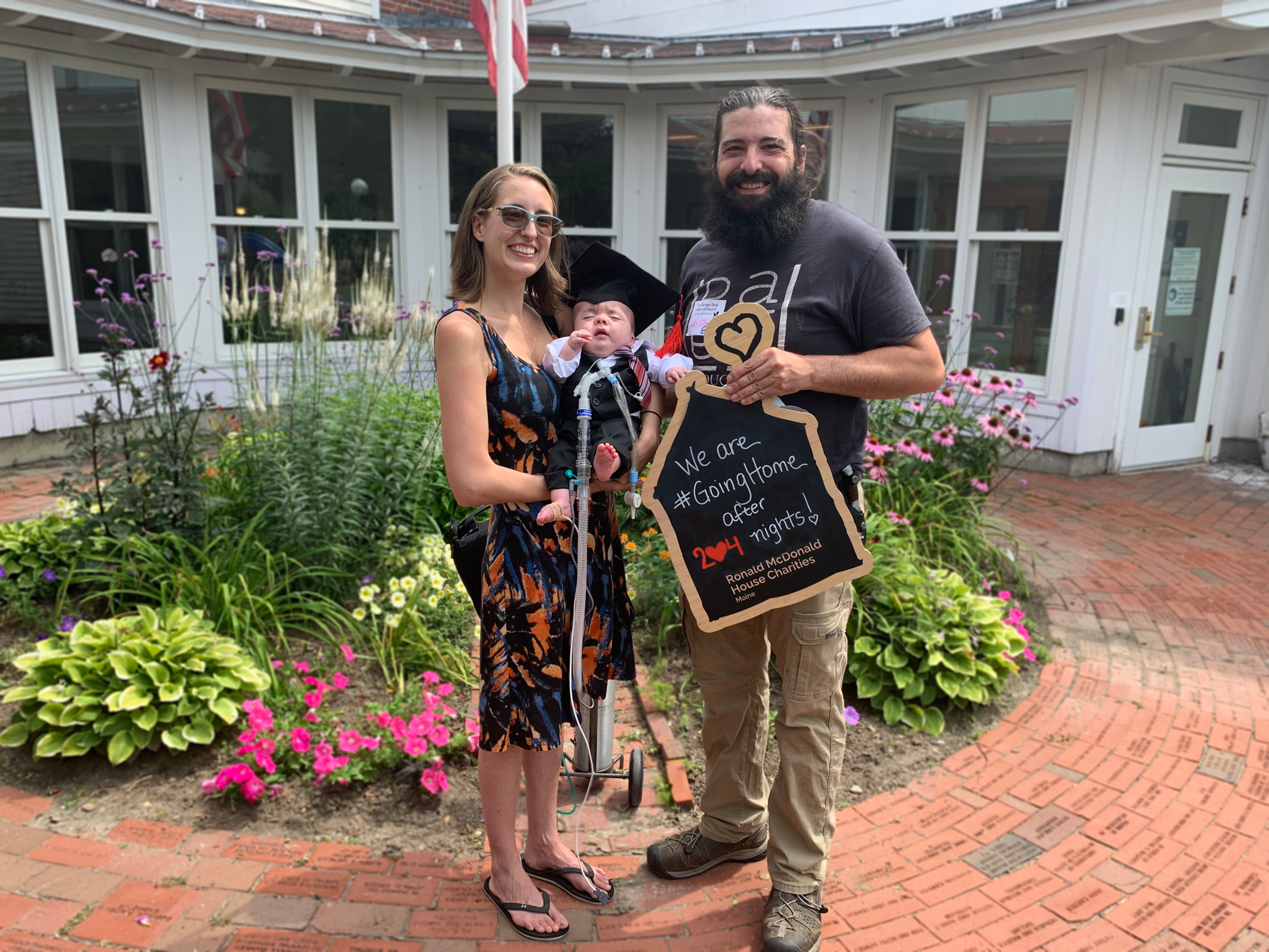 Family poses outside a building with flowers. A baby in graduation attire holds a sign about going home after 204 nights.