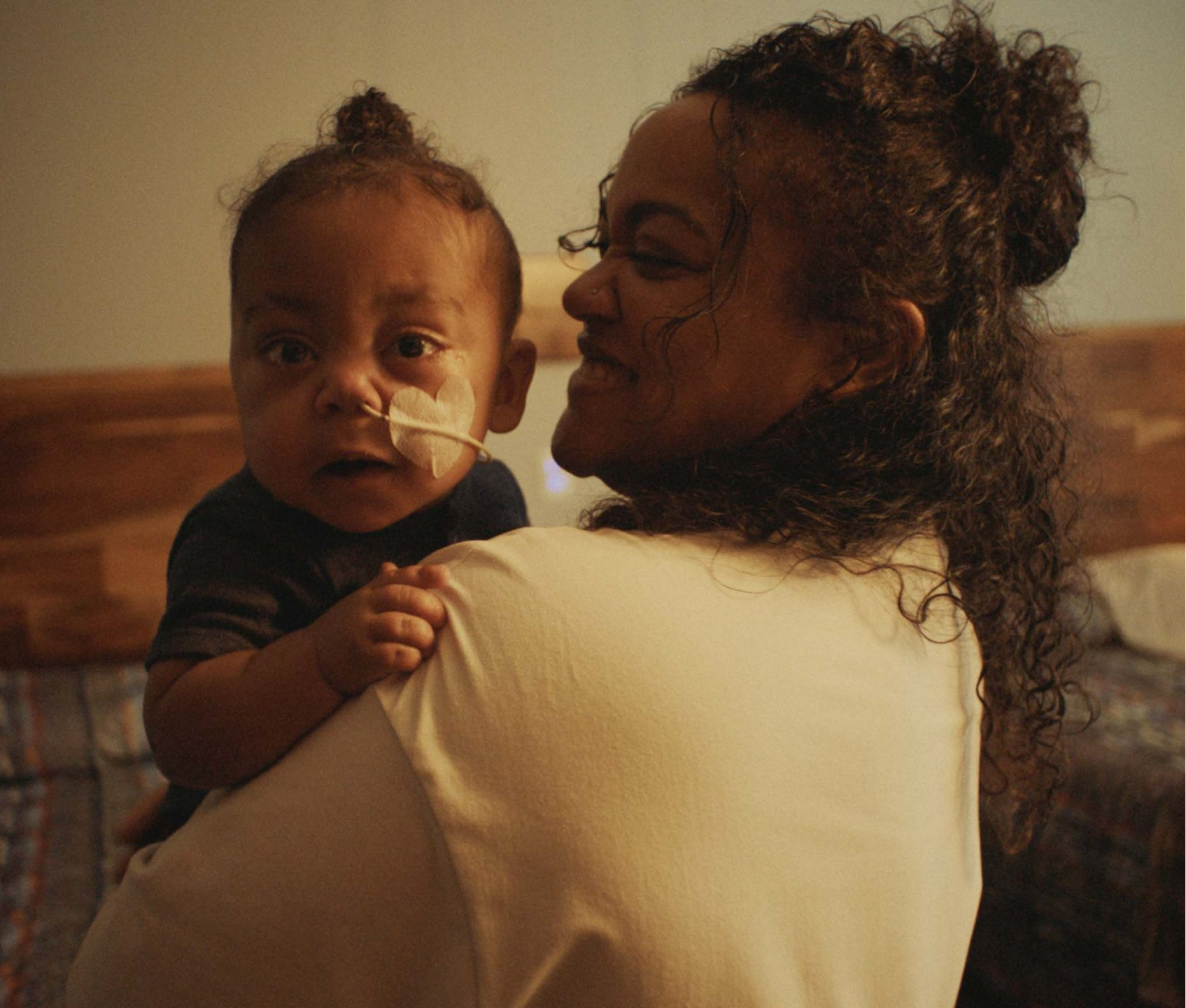 A woman holds a baby with a nasal tube, both smiling warmly at each other in a cozy, softly lit room.