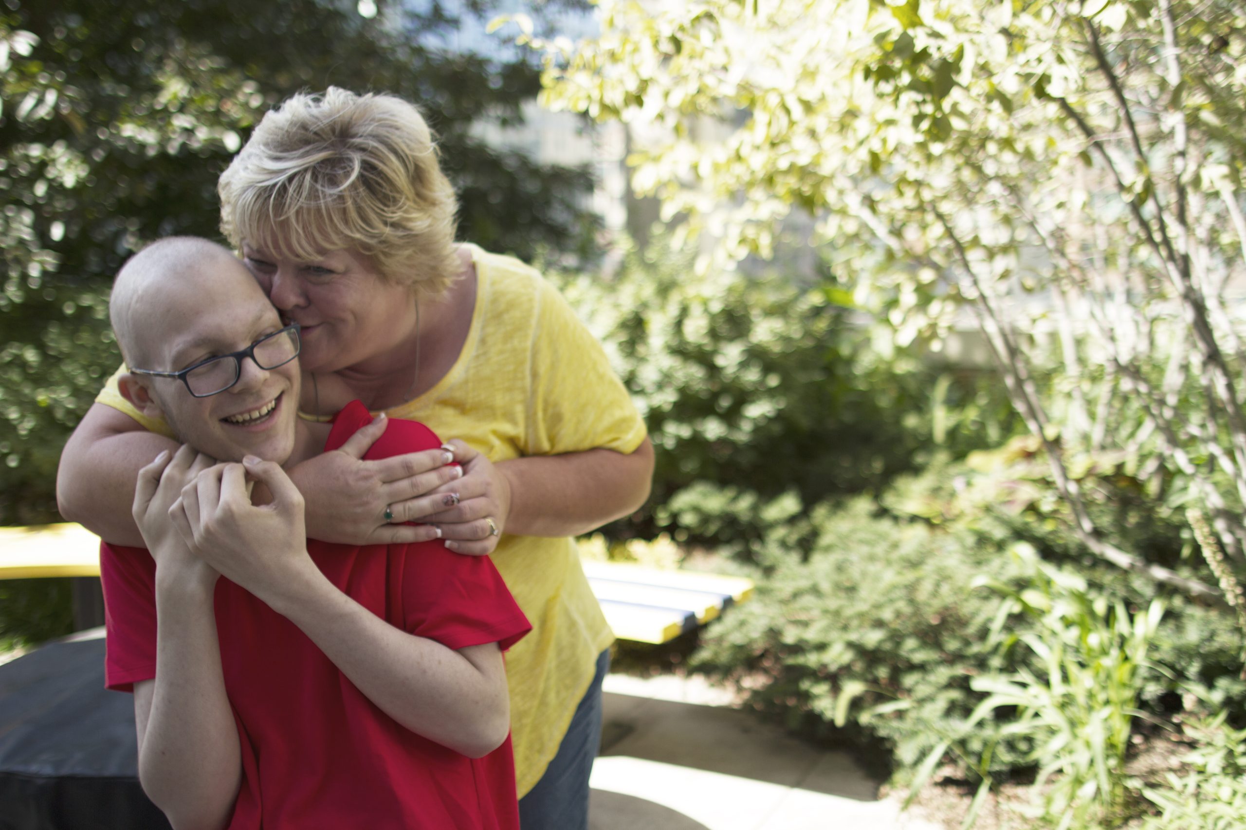 A smiling young man in a red shirt is embraced by a woman in a yellow shirt, surrounded by greenery.
