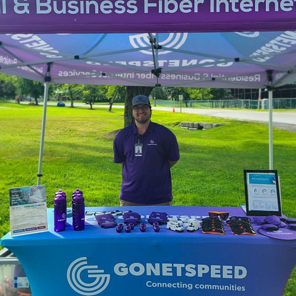 A vendor at a fiber internet booth, smiling, with promotional items and a tablet under a purple canopy in a park.