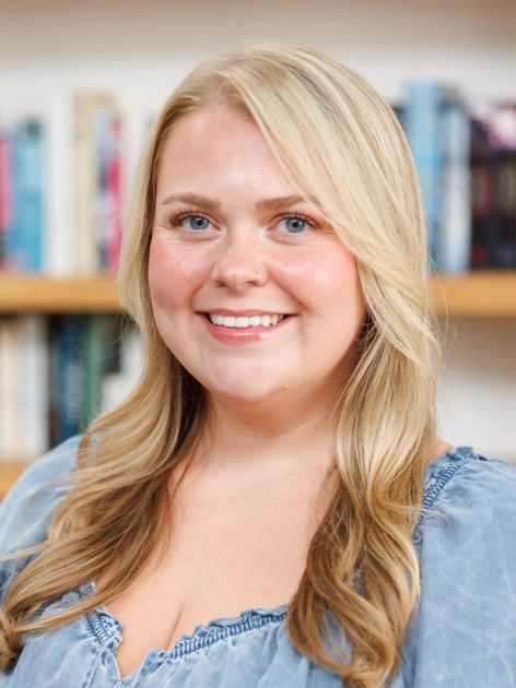 A smiling woman with long, wavy blonde hair, wearing a denim top, in front of a bookshelf.
