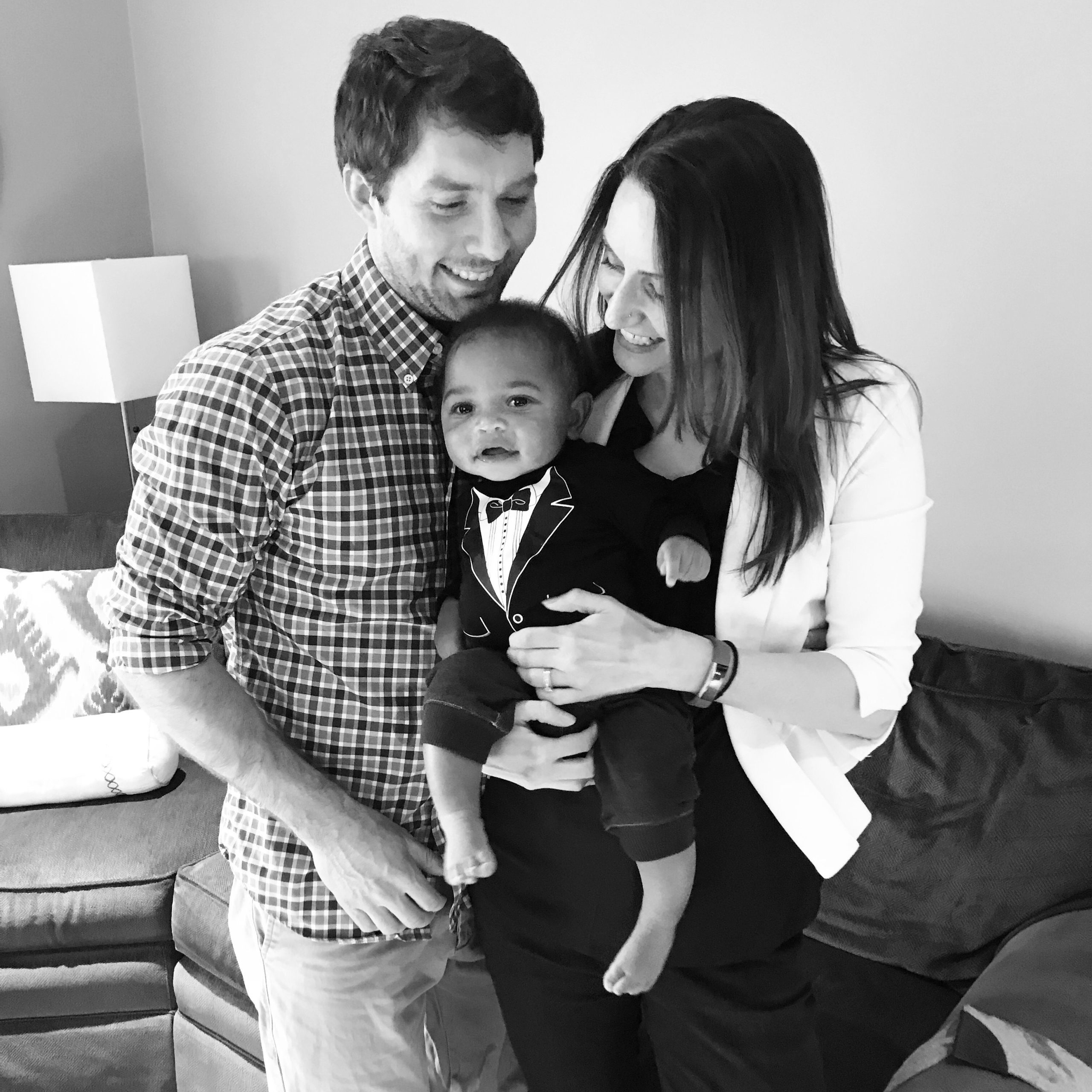 A couple smiles as they hold a baby dressed in a tuxedo, in a cozy living room setting. Black and white photo.