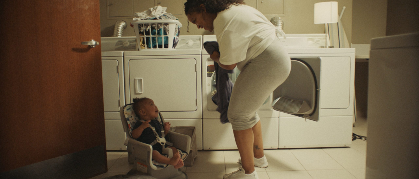 A woman in activewear smiles at a baby in a car seat while doing laundry in a bright, spacious laundry room.