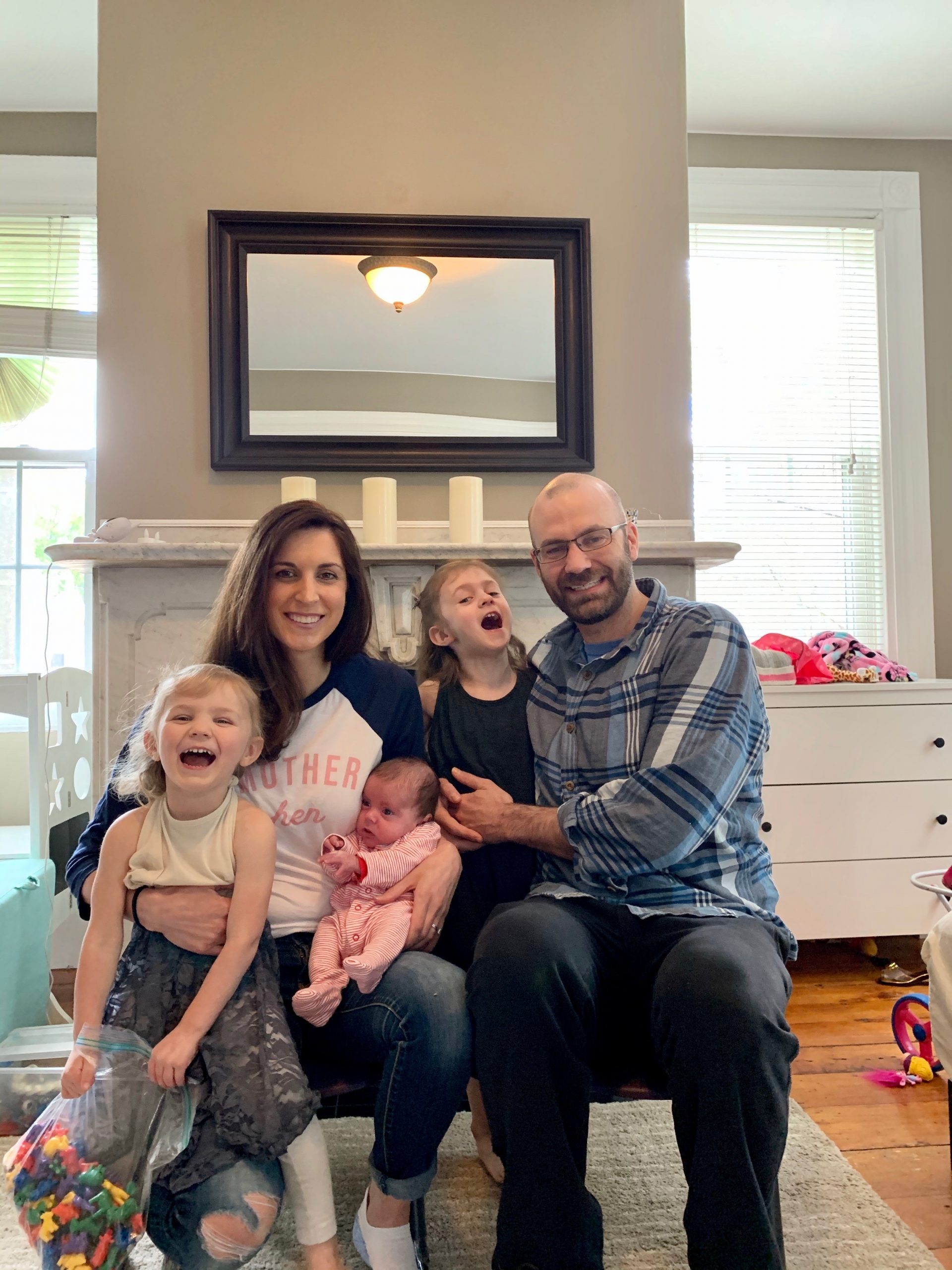 A smiling family of four sits together on a couch, with kids holding colorful toys and a baby in mother's arms.