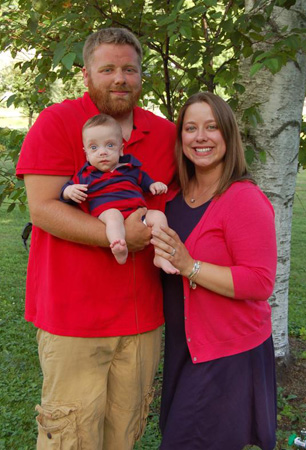 A smiling couple stands together outdoors, holding their baby in a striped shirt, surrounded by green trees.