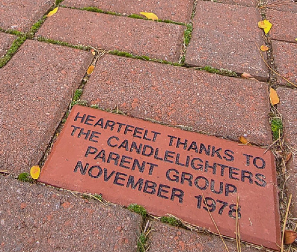 A brick engraved with "Heartfelt thanks to the Candlelighters Parent Group, November 1978," surrounded by pavement and leaves.