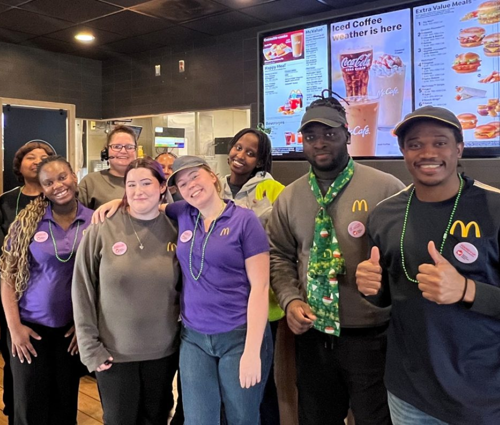 A group of McDonald's employees smiling and posing together in a restaurant, with menu boards visible in the background.