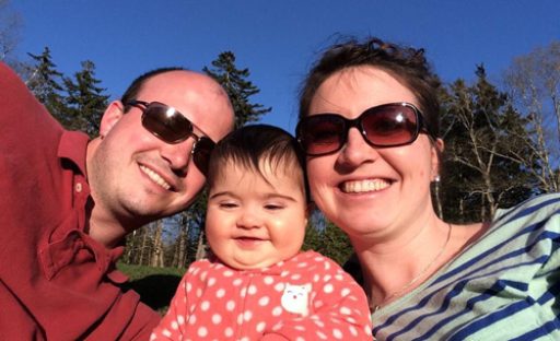 A smiling couple in sunglasses with a baby in a polka-dot onesie, enjoying a sunny day outdoors.