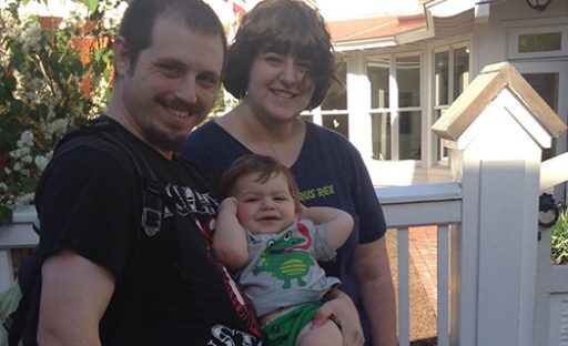 A smiling family poses outdoors, with a man and woman holding a happy baby between them. Bright, sunny day.