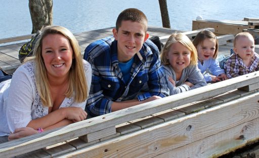 A group of five smiling children and teens lay on a dock by the water, enjoying a sunny day outdoors.