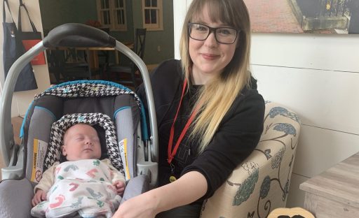 A smiling woman in glasses sits next to a baby in a car seat, wrapped in a blanket, in a cozy indoor setting.