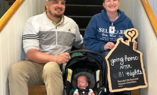 A family sits on stairs with a sign reading "going home after 81 nights" next to a baby in a car seat.