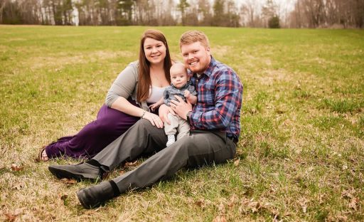 A smiling couple sits on grass, holding a baby, with trees in the background and a peaceful outdoor setting.
