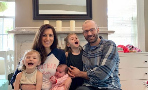 A smiling family of four sits together on a couch, with kids holding colorful toys and a baby in mother's arms.