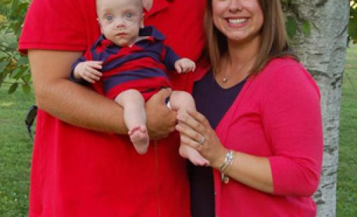 A smiling couple stands together outdoors, holding their baby in a striped shirt, surrounded by green trees.