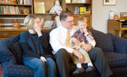 A family sits together on a couch, smiling and interacting in a cozy living room with bookshelves in the background.