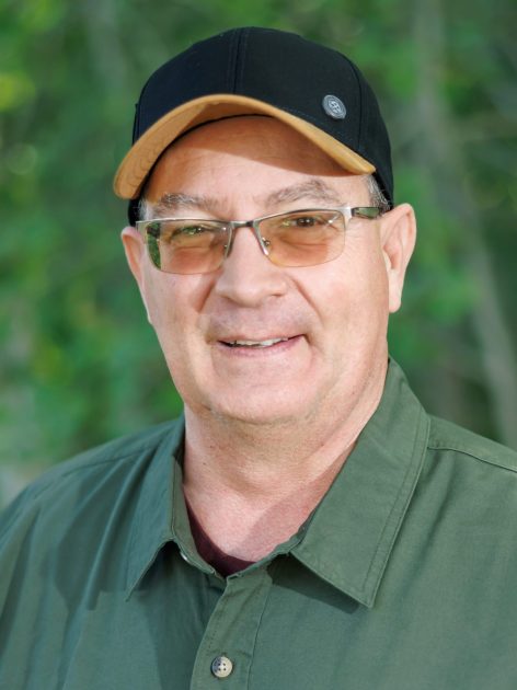 Smiling man in glasses and a cap, wearing a green shirt, with greenery in the background.