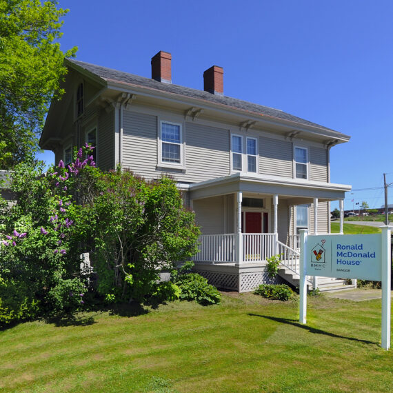Two-story house with a sign for Ronald McDonald House, surrounded by greenery and flowers on a sunny day.