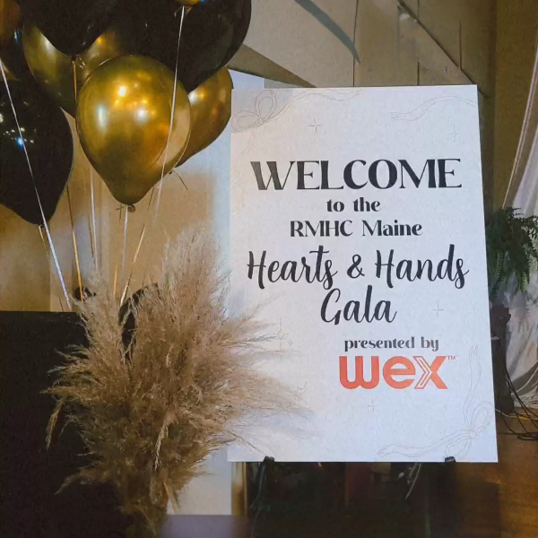 Welcome sign for Ronald McDonald House Maine Maine Hearts & Hands Gala, with black and gold balloons nearby and decorative plants.